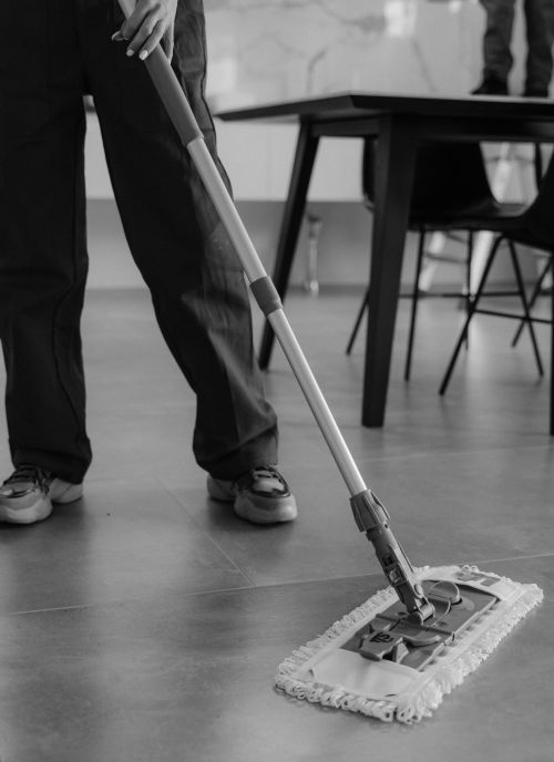 Monochrome image of a person mopping a tiled floor in an indoor setting.